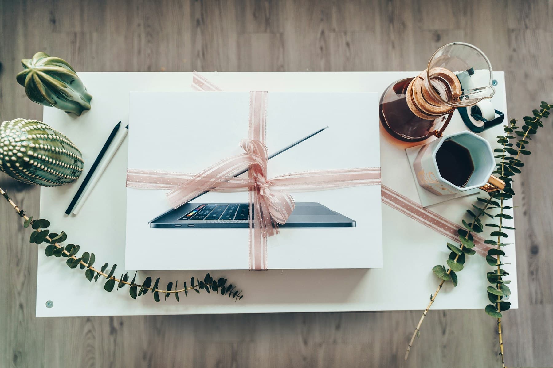 A laptop gift wrapped with a soft pink ribbon beside coffee and greenery